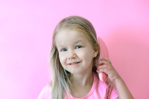 Niña feliz peinándose. Concepto de belleza e infancia. Chica sobre un fondo rosa aislado peinando su cabello con un peine rosa su cabello rubio