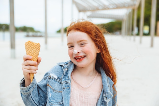Una niña feliz con el pelo rojo come helado en la playa en las vacaciones de verano