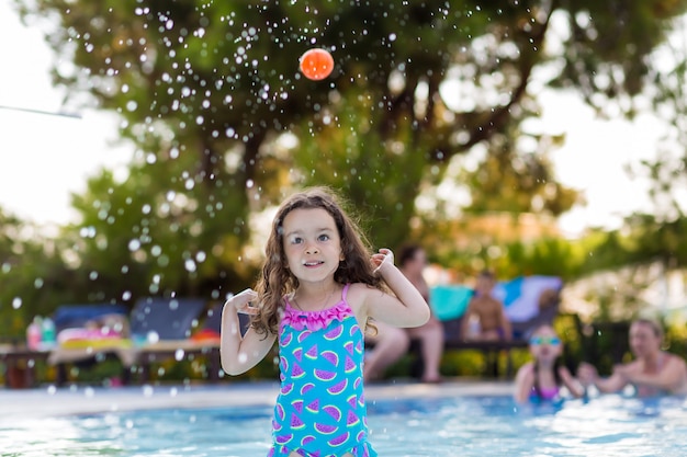 Niña feliz con el pelo suelto en un brillante traje de baño jugando a la pelota en la piscina en un soleado día de verano