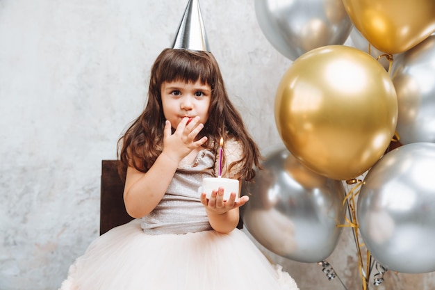 Una niña feliz pide un deseo y sopla las velas de los globos de la torta Espacio para texto cumpleaños decorativo en la pared