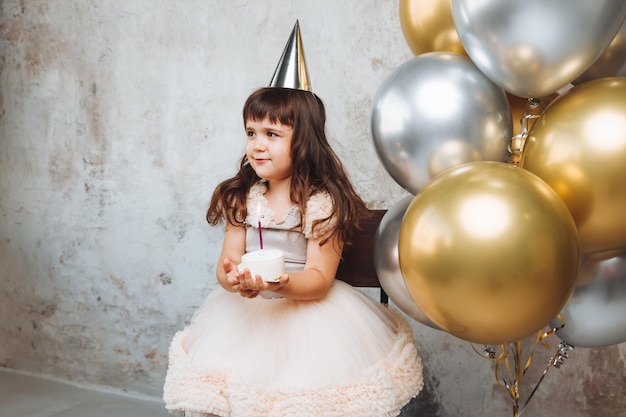 Una niña feliz pide un deseo y sopla las velas de los globos de la torta Espacio para texto cumpleaños decorativo en la pared