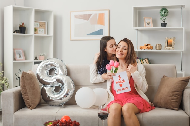 Niña feliz de pie detrás del sofá sosteniendo flores con tarjeta de felicitación abrazó y besando a la madre en el sofá en el día de la mujer feliz en la sala de estar