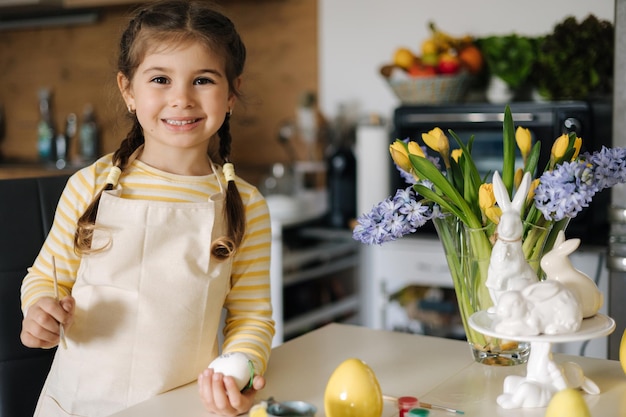 Niña feliz pintando huevos de pascua en la cocina Linda niña sonriente diviértete en las vacaciones de primavera