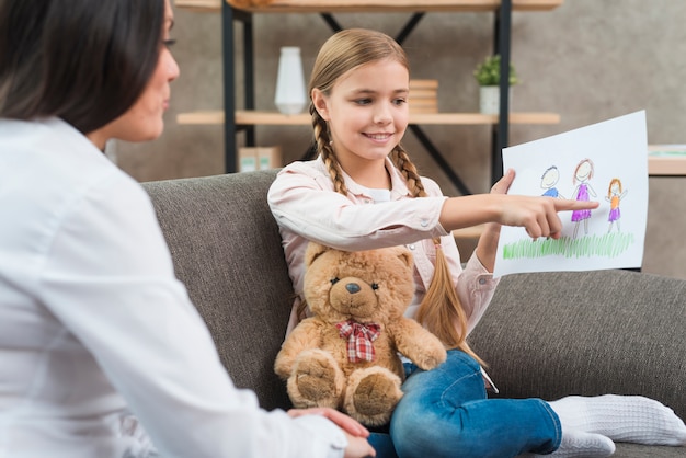 Niña feliz que muestra el dibujo de la familia en papel a la psicóloga