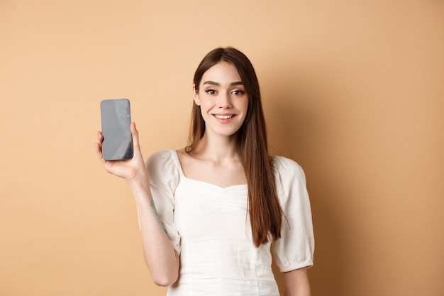 Niña feliz que muestra la pantalla del móvil vacía y sonriendo, demuestra la aplicación del teléfono, de pie sobre fondo beige.