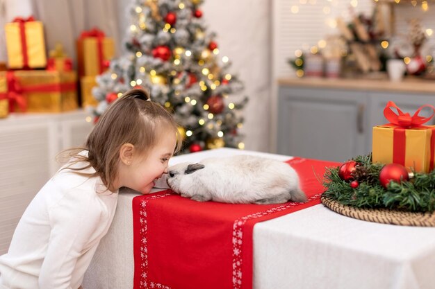 Foto una niña feliz se ríe y se frota la nariz con un conejito en el fondo de un árbol de navidad con luces borrosas