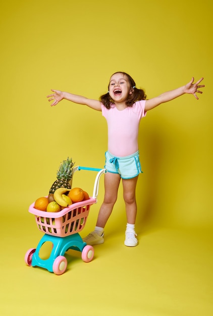 Niña feliz, riendo con los brazos abiertos, de pie sobre un amarillo cerca de su carrito de la compra lleno de deliciosas frutas exóticas.