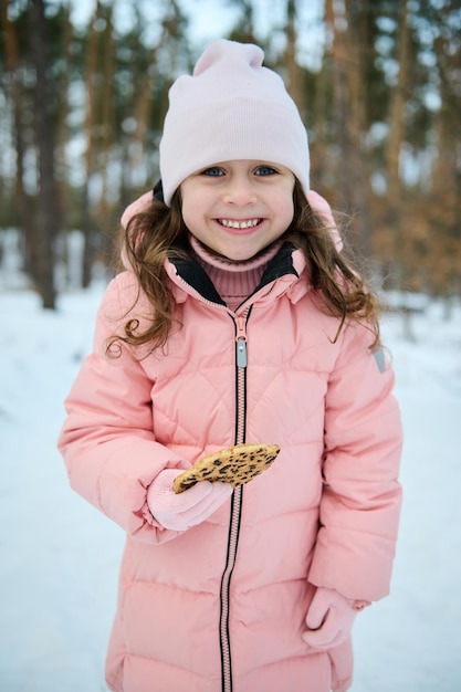 Una niña feliz con ropa de invierno cálida rosa pastel sostiene galletas en la mano y sonríe con una sonrisa dentuda mira a la cámara mientras juega en un bosque nevado Disfruta del maravilloso invierno al aire libre