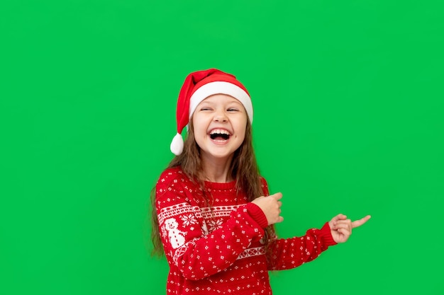 Una niña feliz con ropa de invierno, un sombrero de Papá Noel y un suéter rojo en un fondo verde aislado, muestra sus dedos en algo y sonríe en un lugar o espacio para texto