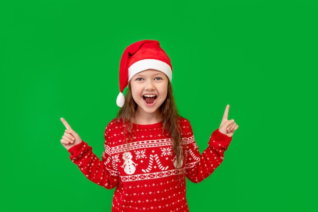 Una niña feliz con ropa de invierno, un sombrero de Papá Noel y un suéter rojo en un fondo verde aislado, muestra sus dedos en algo y sonríe en un lugar o espacio para texto