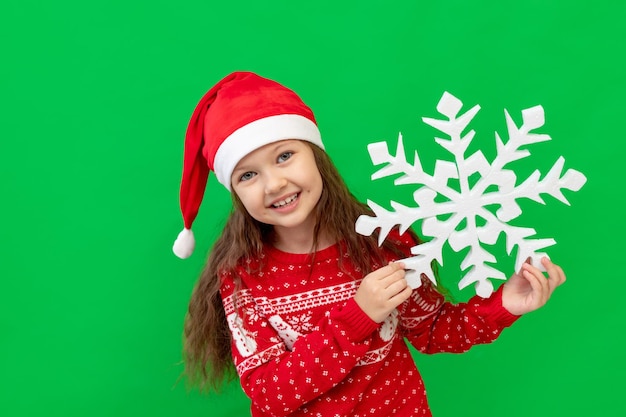 Una niña feliz en ropa de invierno, sombrero de Santa Claus y suéter rojo sobre un fondo verde aislado tiene un gran copo de nieve y sonrisas, un lugar o espacio para texto