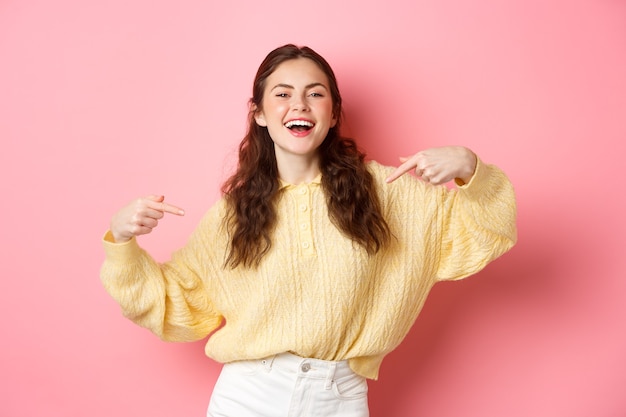 La niña feliz segura de sí misma se señala a sí misma y se ríe, promocionándose a sí misma y sonriendo, de pie contra la pared rosa.