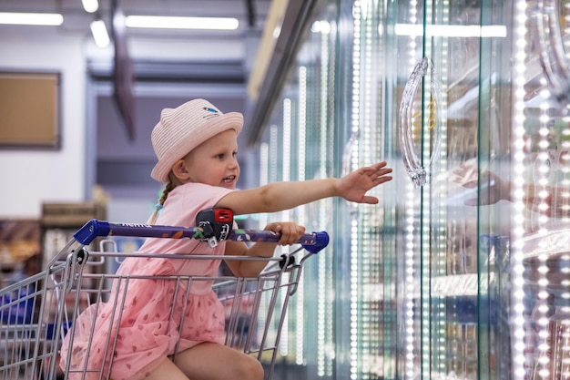 Una niña feliz sentada en un carrito de compras en una tienda de alimentos extiende su mano