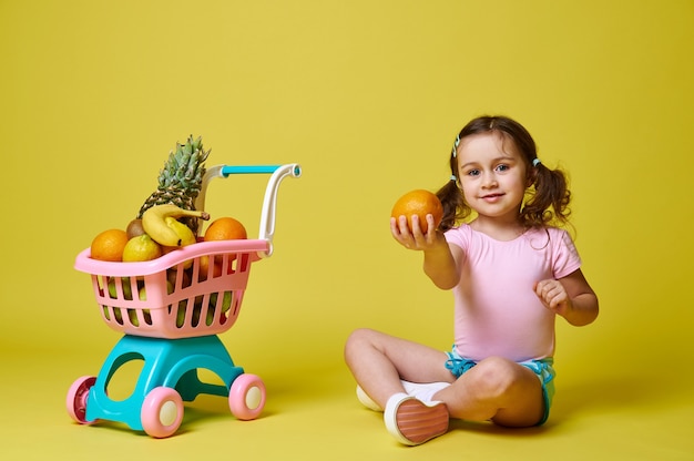 Niña feliz sentada cerca de un carrito de compras lleno de frutas sobre una superficie amarilla, sosteniendo una naranja y mostrándola a la cámara. Aislado en amarillo con espacio de copia