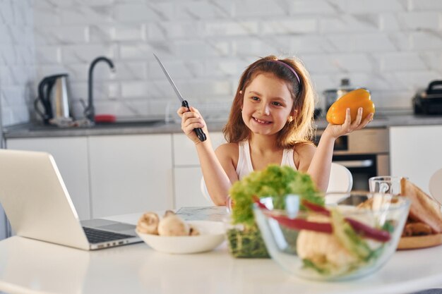 Una niña feliz se sienta en la cocina con comida y una laptop en la mesa