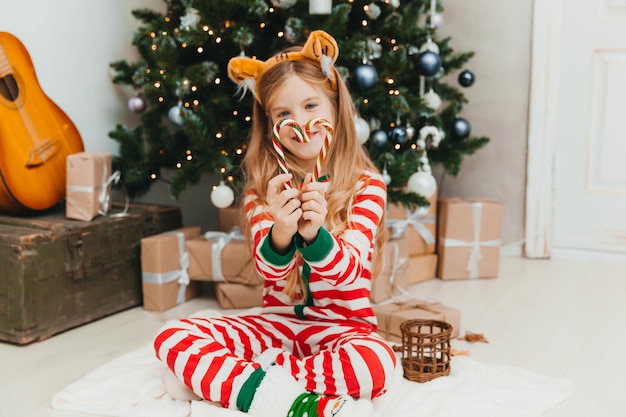 Niña feliz se sienta con dulces cerca del árbol de Navidad. Navidad.