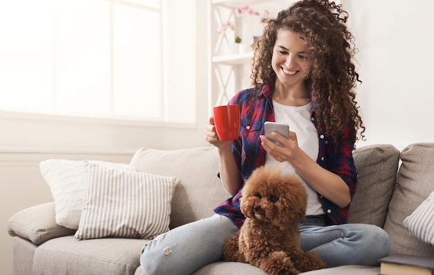 Niña feliz con smartphone y perro en casa. Mensajería de mujer rizada en línea en el sofá con su cachorro, espacio de copia