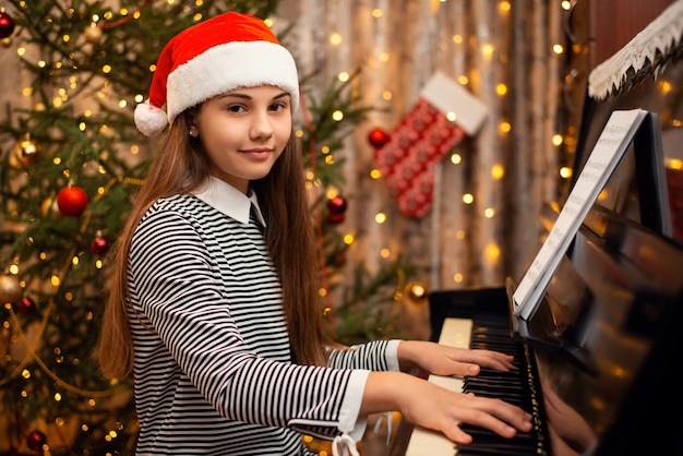 Niña feliz con sombrero de Navidad sentado cerca de abeto decorado y tocando el piano. Año nuevo, vacaciones, tiempo en casa con concepto de familia.