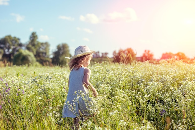 Niña feliz con sombrero de paja caminando en el campo de flores en un día soleado de verano