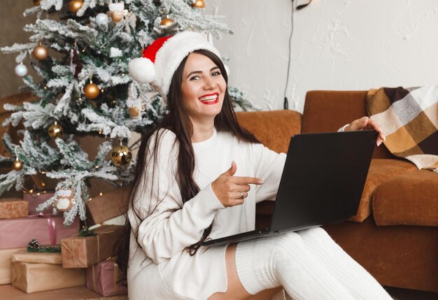 Una niña feliz con un sombrero de Papá Noel desea a sus amigos Feliz Navidad en un chat de video en una computadora portátil Una mujer joven acostada debajo de un árbol de Navidad entre cajas de regalo en el interior de una casa