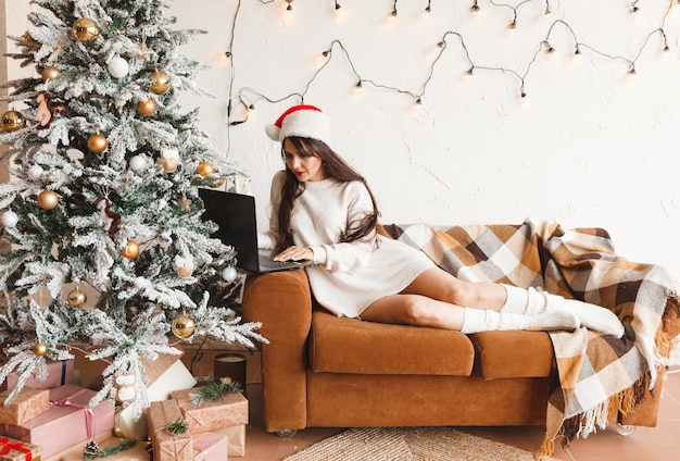 Una niña feliz con un sombrero de Papá Noel desea a sus amigos Feliz Navidad en un chat de video en una computadora portátil Una mujer joven acostada debajo de un árbol de Navidad entre cajas de regalo en el interior de una casa