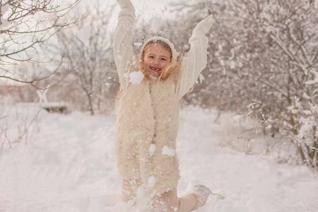 una niña feliz con un sombrero de punto blanco y un suéter está jugando bolas de nieve en un parque nevado