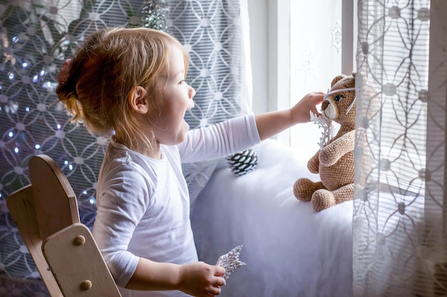 niña feliz sonriendo jugando con copos de nieve hechos a mano y oso de peluche de juguete cerca de la ventana de luz