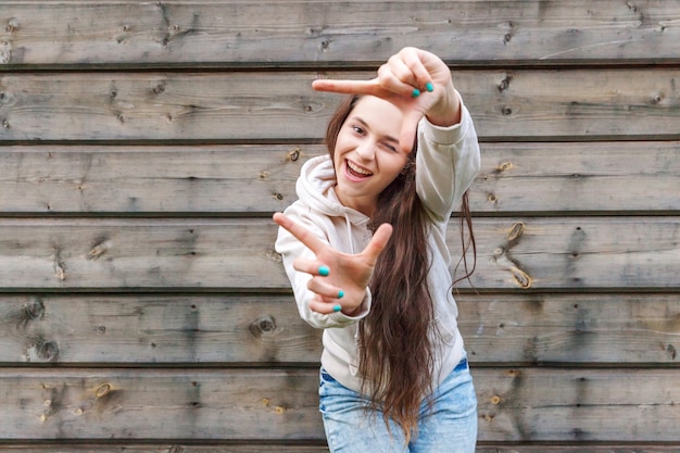 Niña feliz sonriendo. Retrato de belleza joven mujer morena riendo mostrando el marco con los dedos sobre fondo de pared de madera. Mujer europea. Expresión facial de emociones humanas positivas, lenguaje corporal