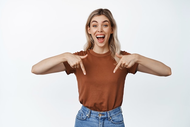 Niña feliz sonriendo, señalando con el dedo hacia abajo con expresión entusiasta, invitando a ver el evento, mostrando algo, de pie en blanco