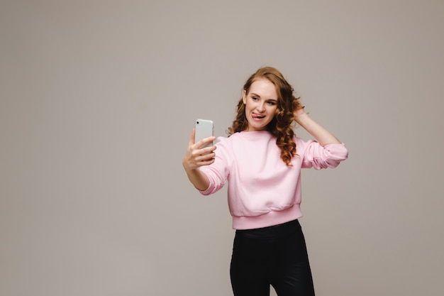 Una niña feliz sonriente con una blusa rosa toma un selfie en un teléfono inteligente en un gris.