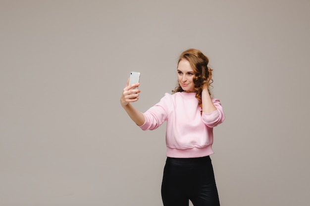 Una niña feliz sonriente con una blusa rosa toma un selfie en un teléfono inteligente sobre un fondo gris.