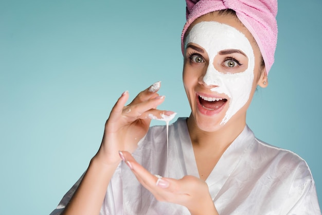 Niña feliz sorprendida con una toalla rosa en la cabeza pone una mascarilla nutritiva blanca en su rostro