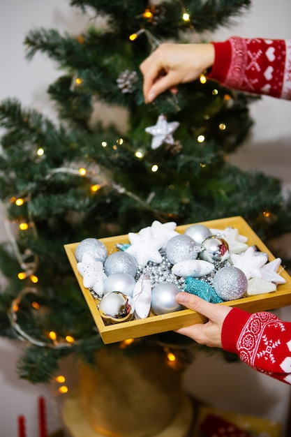 Niña feliz sostiene una bandeja con juguetes navideños en sus manos y decora el árbol de Navidad en casa para Navidad, año 2022 del tigre.