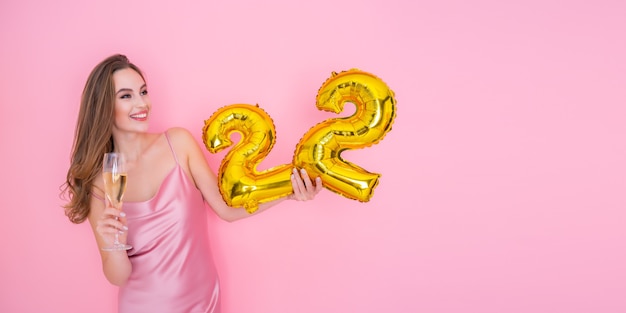 Niña feliz sostiene un globo de lámina de oro y una copa de champán en la fiesta de cumpleaños de fondo rosa