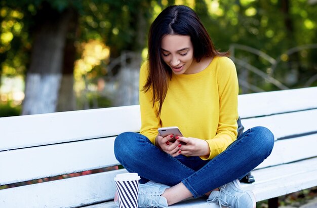 Una niña feliz sostiene en las manos el teléfono inteligente durante la compra en las ventas Una mujer joven tiene una expresión alegre durante la conversación en línea con sus amigos en las redes sociales sentada en el banco del parque