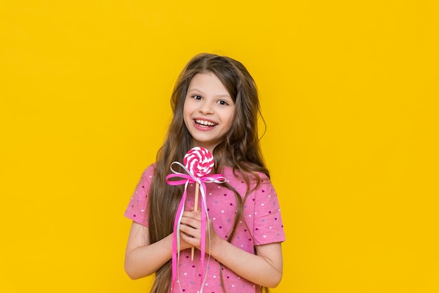 Foto una niña feliz sostiene una piruleta de caramelo en sus manos y sonríe a una niña con un caramelo en un fondo amarillo aislado