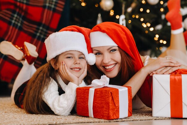 Una niña feliz y su mamá con sombreros de Santa Claus están sonriendo con regalos en sus manos.