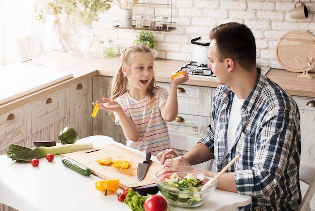Niña feliz y su papá cocinando en la cocina de casa. Padre e hija haciendo ensalada de verduras y divirtiéndose juntos. Día del padre, familia y concepto de comida sana.