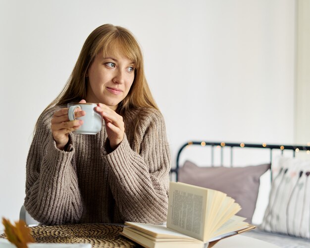 Niña feliz en suéter soñando con una taza de té mientras lee un libro en el dormitorio