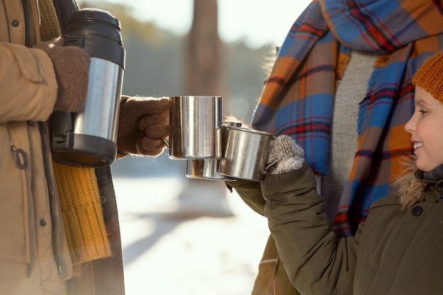 Niña feliz y sus padres en ropa de invierno abrigada tintineando con tazas metálicas con té caliente mientras se calienta afuera en un día helado de invierno