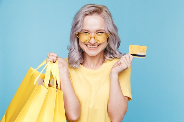 Niña feliz con tarjeta de crédito y grandes bolsas de compras aisladas sobre el estudio azul