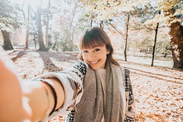 Niña feliz con teléfono inteligente durante el otoño en un parque tomando selfie, mujer hablando por móvil en otoño. Videollamada al aire libre, recibiendo buenas noticias.