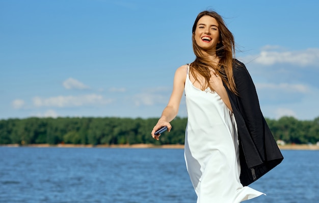 Niña feliz con teléfono en vestido blanco con chaqueta junto al mar. Foto de alta calidad