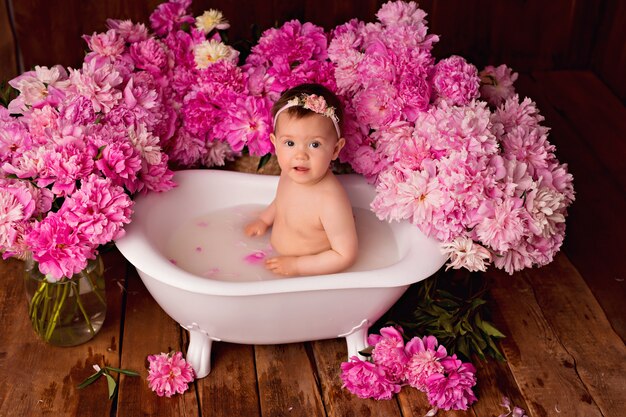 Niña feliz toma un baño de leche con pétalos. Ramos de peonías rosas.