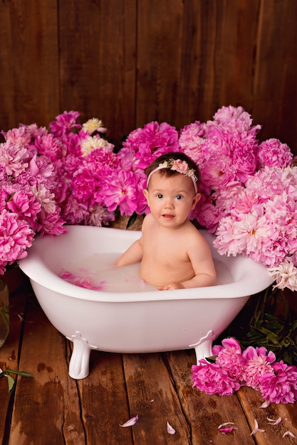 Niña feliz toma un baño de leche con pétalos. Ramos de peonías rosas.