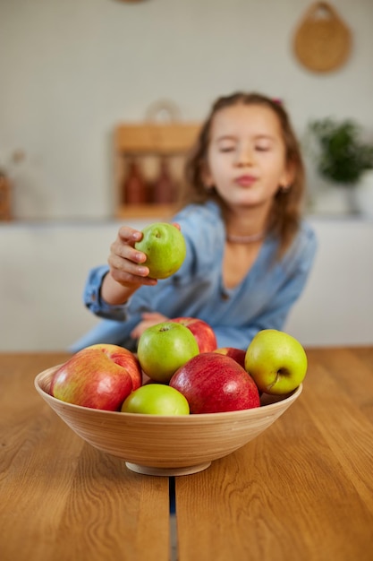 Niña feliz toma una pera del tazón con variedad de frutas en la mesa en casa niño comiendo bocadillos saludables nutrición vegetariana para niños vitaminas para niños