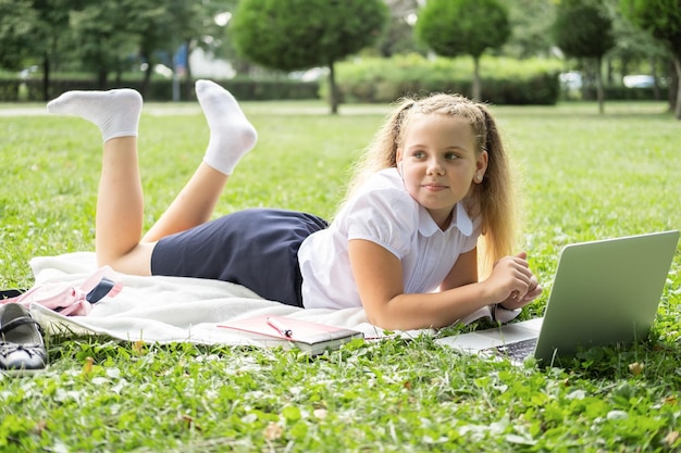 niña feliz en uniforme escolar con cuaderno tiene lección en línea en la computadora portátil en el césped