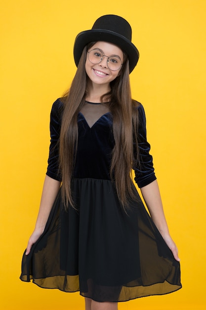 Niña feliz usa vestido de fiesta negro y sombrero en el pelo largo morena con fondo amarillo de aspecto de belleza, salón.