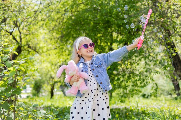 Una niña feliz en verano con gafas de sol infla pompas de jabón tiene un juguete en sus manos