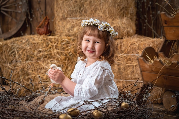 Niña feliz con un vestido y corona se sienta en un nido y sostiene un lindo pollo.